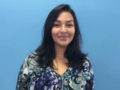 A person is smiling while standing against a plain blue background, wearing a floral-patterned shirt.