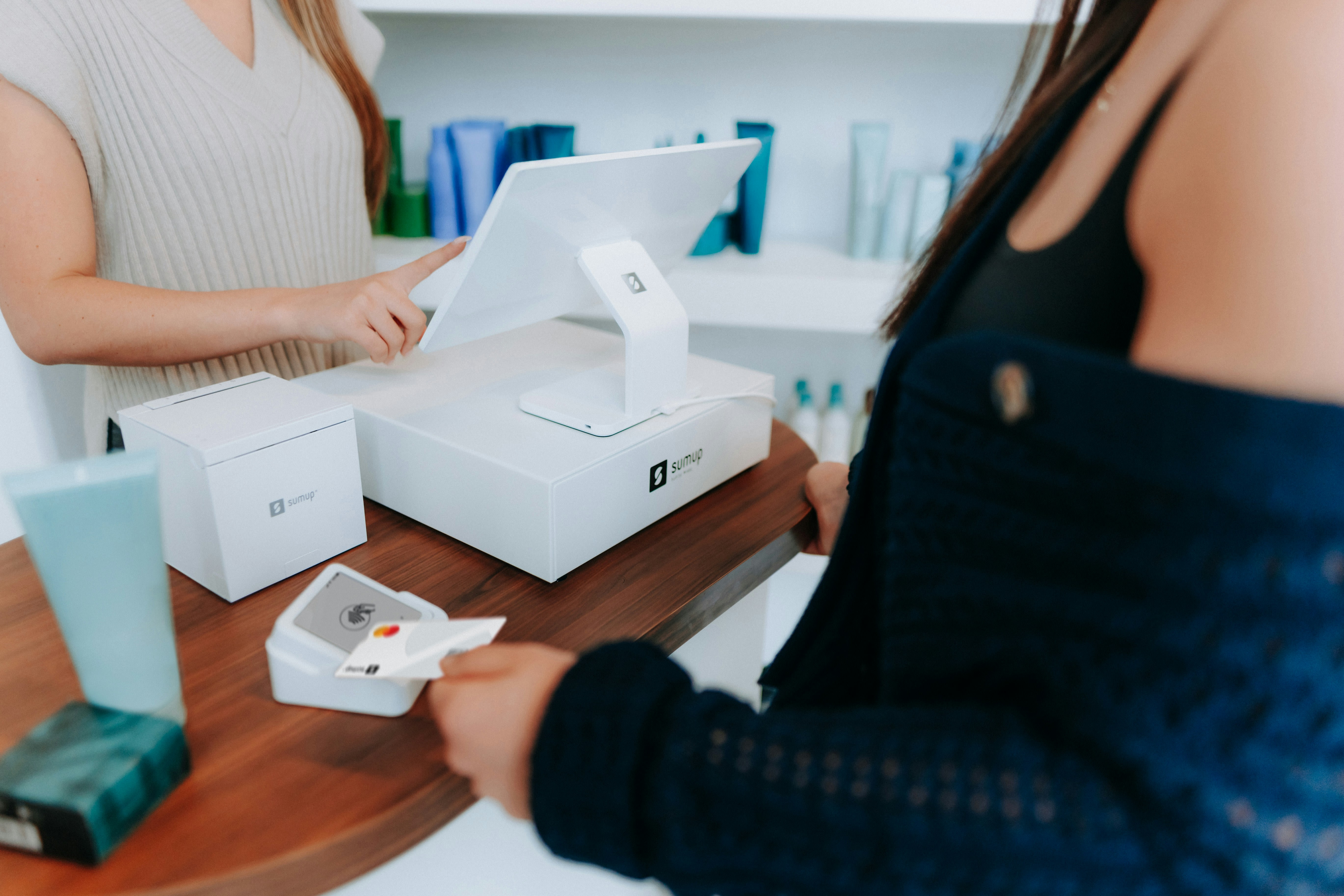 A person uses a SumUp card reader to make a payment. Another person operates a touchscreen device labeled "sumup" on a counter, with colorful bottles in the background.