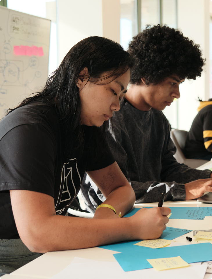Two SumUp team members focused on writing ideas on blue and yellow sticky notes during a Hack Week workshop session, with a whiteboard and natural light in the background.