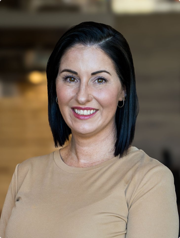 A woman with black hair and a warm smile poses in a modern office. She wears a beige top and gold hoop earrings. The softly blurred background with warm lighting reflects SumUp’s professional and inclusive workplace in the USA