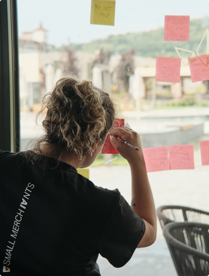 Female SumUp team member wearing a 'Small Merchants' t-shirt writing on a pink sticky note attached to a glass window, with multiple colourful notes and an outdoor view in the background, during a Hack Week brainstorming session.
