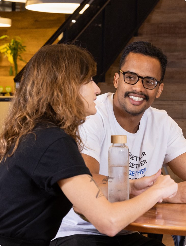 A man wearing glasses and a white t-shirt smiles while engaging in conversation with a woman with wavy brown hair at a wooden table. A reusable water bottle sits in front of them. The setting is SumUp's modern, warmly lit office in São Paulo