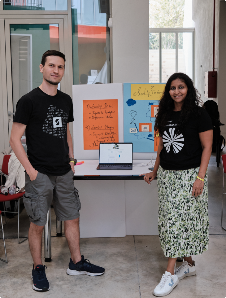 Two SumUp team members standing beside their Hack Week project display, featuring colourful posters, a laptop with a prototype, and handwritten notes, inside a bright open workspace.