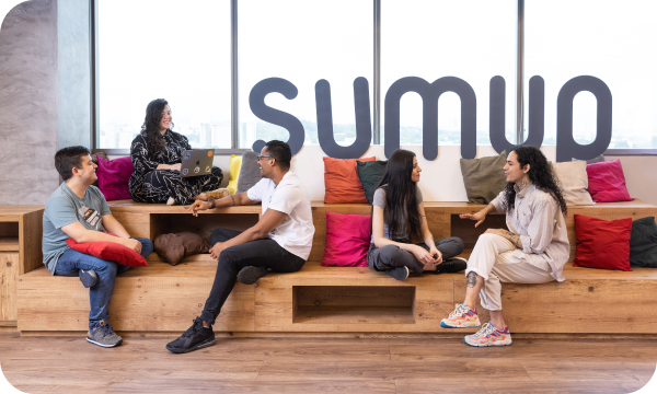 A diverse group of SumUp employees sitting and talking in front of a SumUp sign on the wall