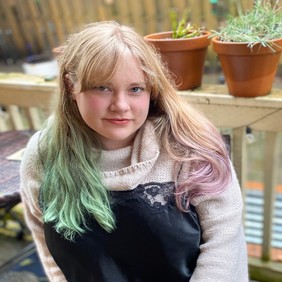 A person with light hair, partly dyed green, sits outdoors on a wooden deck