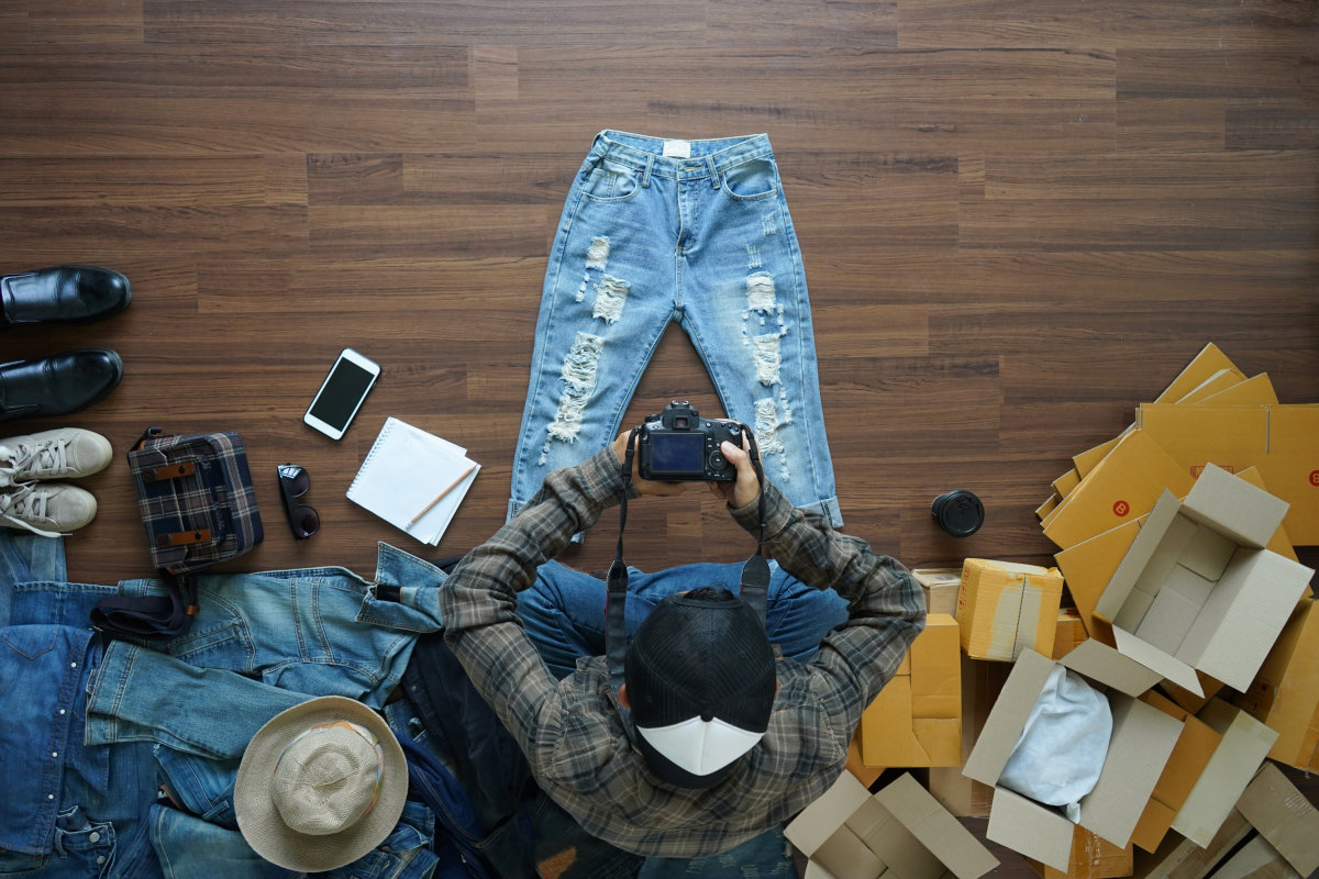 Un uomo seduto a terra scatta una foto di un paio di jeans con una macchina fotografica. I pantaloni sono circondati da altri capi di abbigliamento e imballi vuoti. 