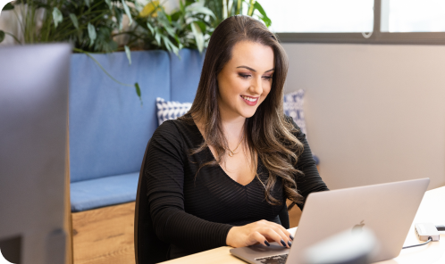 Smiling woman working on a laptop at a desk in a modern office, with green plants and a blue cushioned bench in the background.
