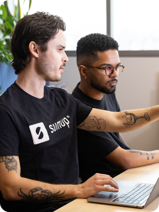 2 male SumUppers wearing black t-shirts, one with the SumUp logo, working together and pointing at a computer.