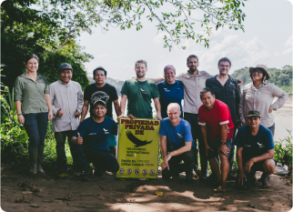 A group from Wilderness International in a green rainforest near a river, holding a yellow sign reading 'Propiedad Privada' with the Wilderness International logo, smiling at the camera. The bright sky has scattered clouds framed by trees