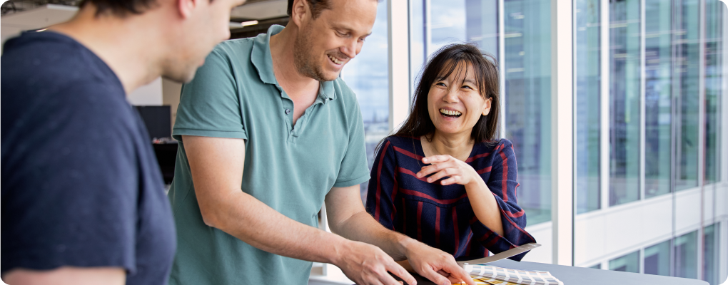 Three colleagues collaborate at a table by large office windows. One person points at a document while another smiles and gestures, creating a positive and engaging atmosphere.