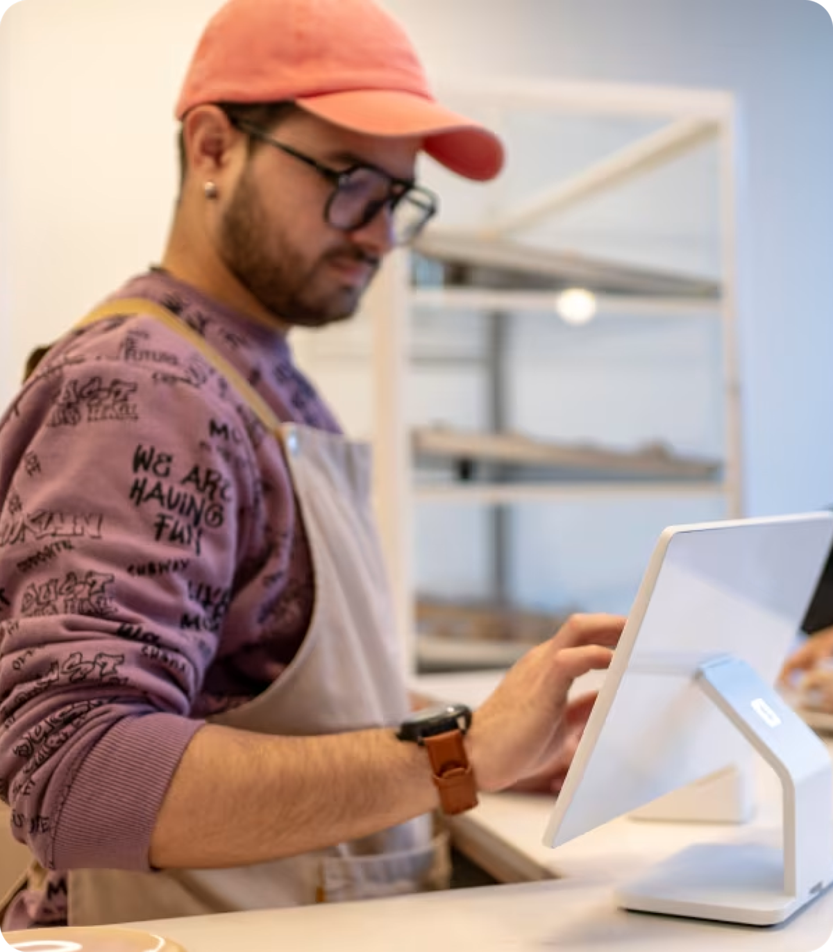 Man wearing a pink hat and apron using a touchscreen point-of-sale terminal in a store or café.