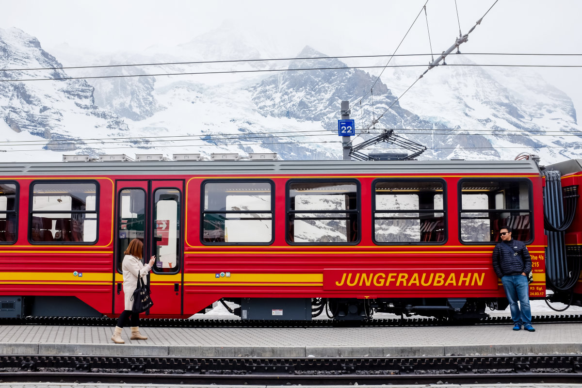 The Jungfraubahn waits at a platform as a woman walks by and a man leans against the train.