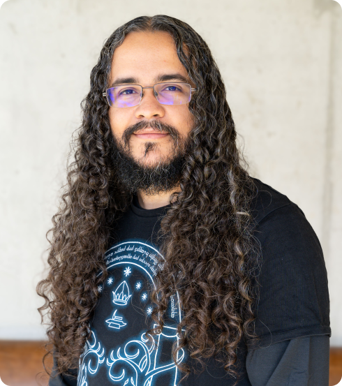 Portrait of a male engineer with long curly hair and a beard, wearing glasses with purple-tinted lenses and a black shirt featuring a glowing blue graphic design, standing against a light-colored background."
