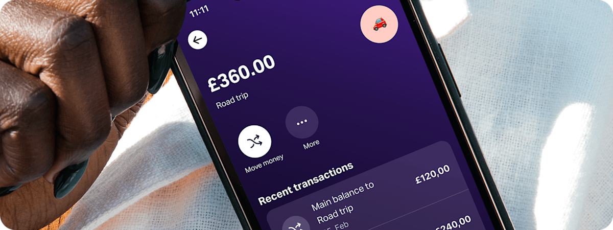 Close-up of a hand holding a smartphone displaying the SumUp Pay app, showing a ‘Road trip’ savings space.
