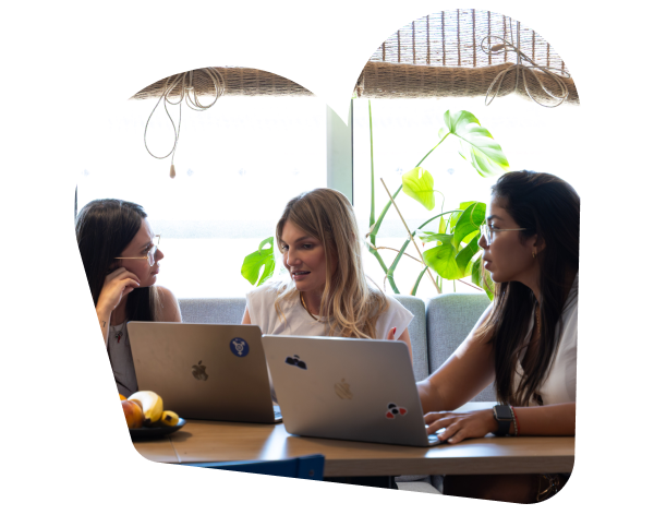 3 women collaborate at a shared table in the SumUp Barcelona office, each with a laptop open. Natural light and lush greenery create a bright.