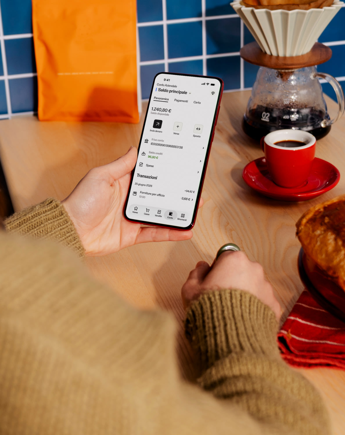 Una persona tiene in mano uno smartphone che mostra l’app SumUp Business, con un caffè e un pasticcino sul tavolo in un ambiente accogliente