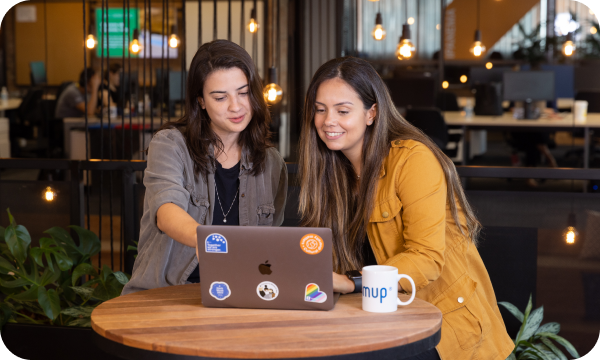 2 female SumUp employees looking and pointing at a laptop covered with colourful stickers and a branded white SumUp mug.