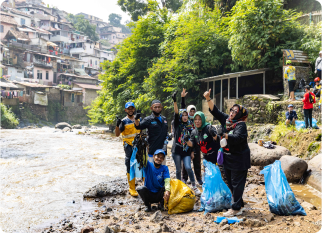 A group of volunteers by a river in Indonesia, holding bags of collected plastic waste. They wear gloves and casual outdoor clothing, smiling and raising their hands in celebration. Behind them, a hillside town with colourful houses and greenery is visible