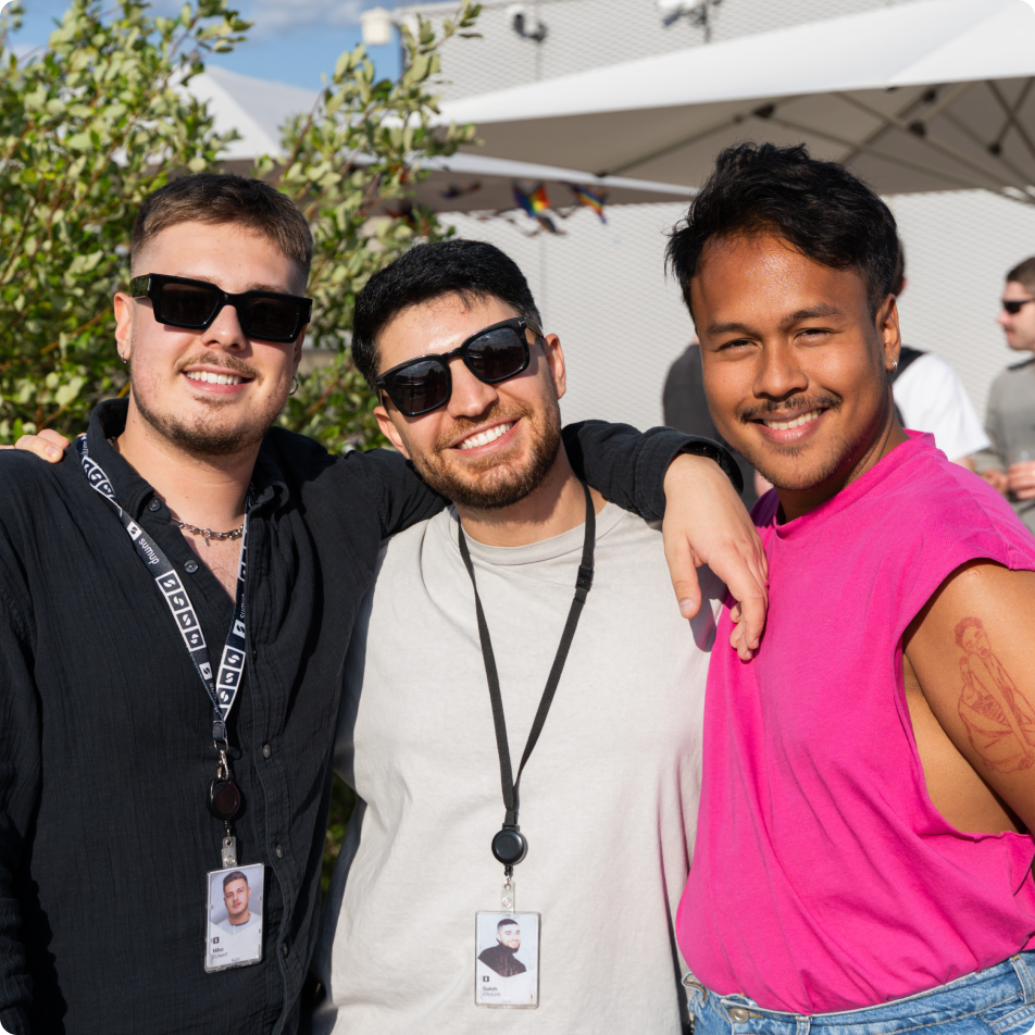 Three SumUppers smiling outdoors on a sunny day with arms around each other during a company Pride Event.