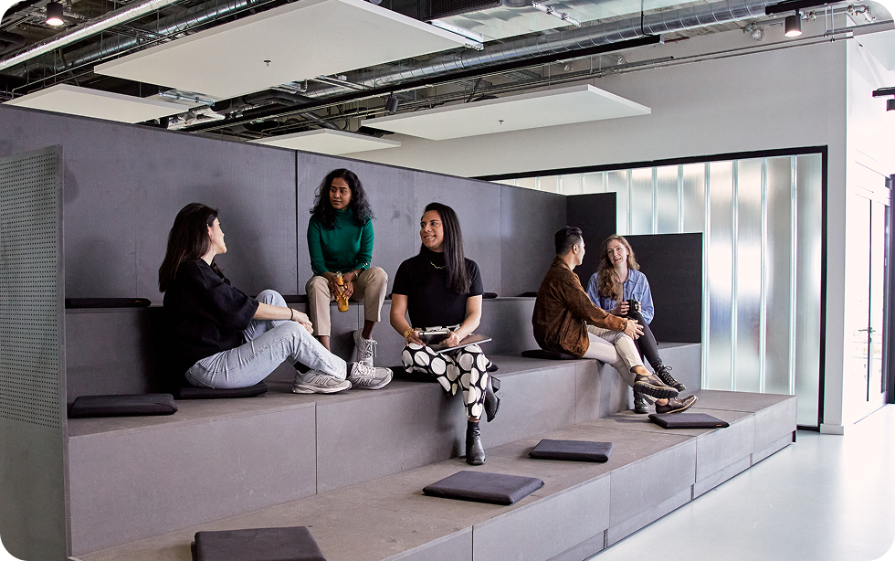 SumUp employees chatting in a modern European office breakout area with tiered seating, gray minimalist design, and industrial ceilings. The relaxed setting highlights collaboration, diversity, and contemporary workspace culture.