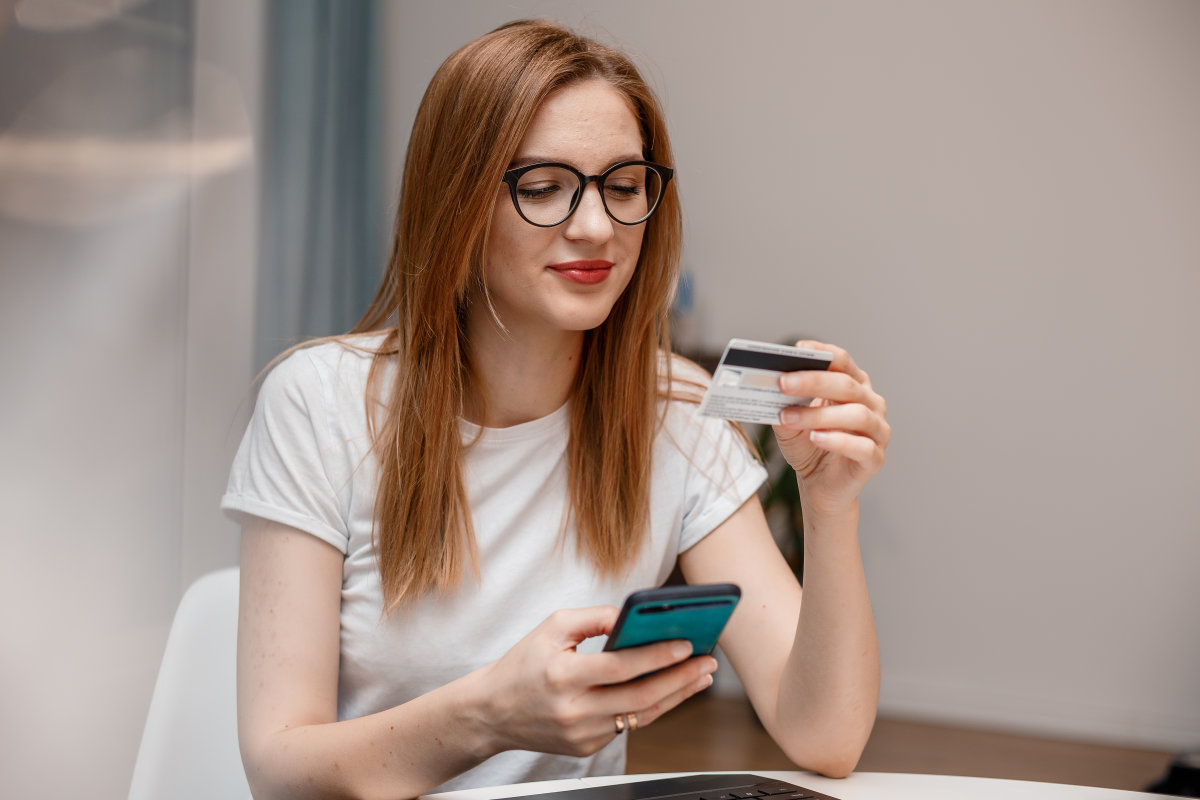 A woman sitting at a table while holding her smartphone and looking at the details on her debit card.