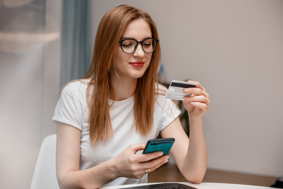 Una donna è seduta a un tavolo con lo smartphone in mano e verifica i dati della sua carta di debito.