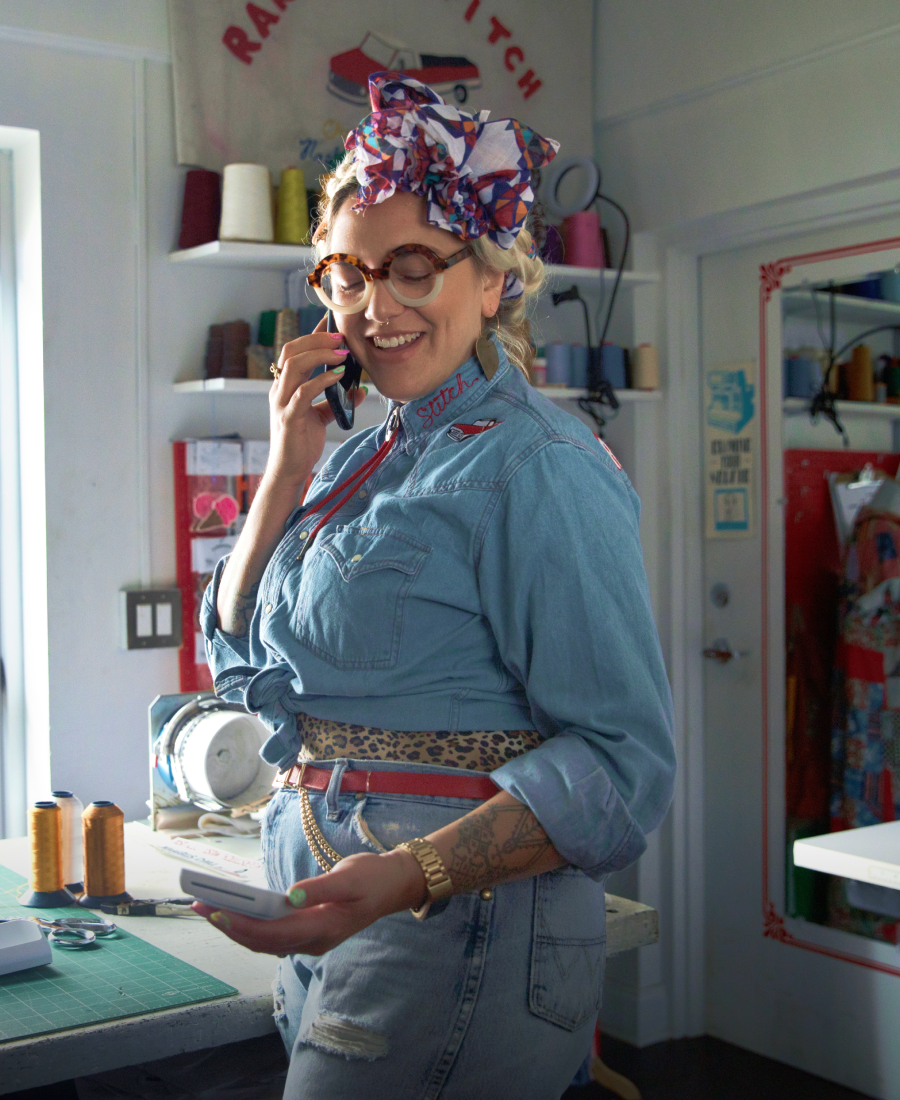 Image showing a person wearing a denim shirt and jeans, with glasses and a colourful hair scarf, holding a SumUp Solo Lite card reader