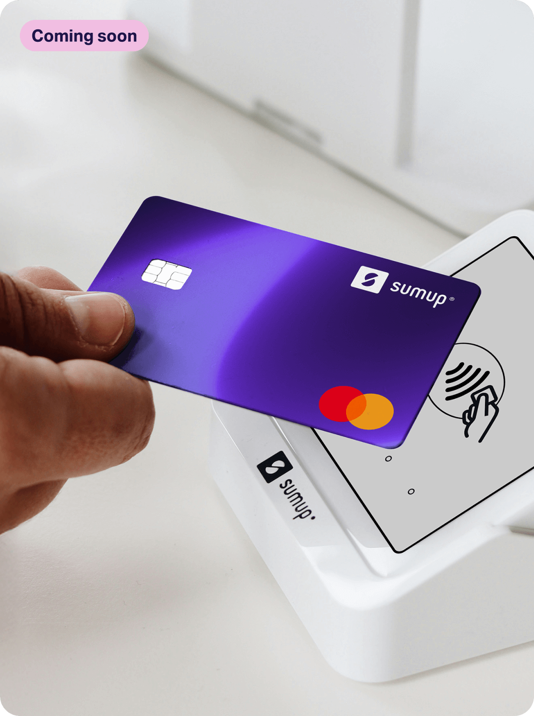 Person holding SumUp Pay's physical debit card near a SumUp card reader for contactless payment. Pink "coming soon" label on the top left.