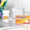 A box of Motrin Migraine pain reliever, a bottle of the same medication, two capsules, a glass of water, and office supplies near a laptop on a desk.