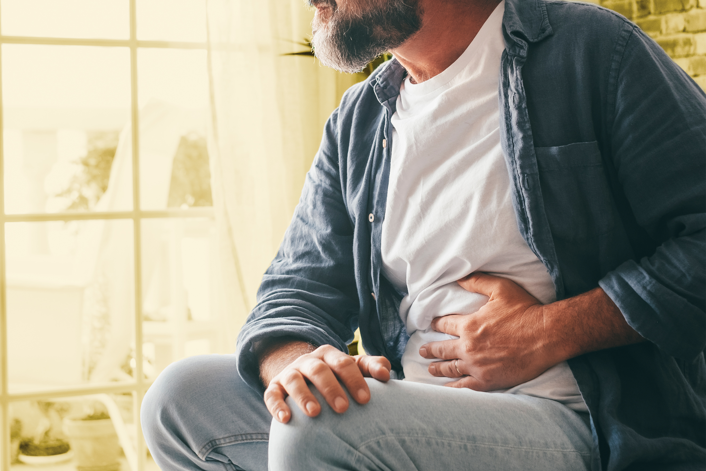 The image depicts a middle-aged man experiencing abdominal discomfort. He is seated, with his knees drawn up towards his chest, and both hands firmly clasped over his stomach area. His expression is not fully visible, but his posture strongly suggests pain or distress. He has a full, dark beard and appears to be wearing a casual outfit consisting of a white t-shirt layered under a dark blue, button-down shirt. He is also wearing light-wash denim jeans and a simple wedding band is visible on his left ring finger. The background is softly blurred, suggesting an indoor setting with a window providing diffused light. The window itself has a textured, geometric pattern, possibly made of frosted glass or a decorative screen. A glimpse of greenery, likely a plant, is visible near the window, adding a touch of life to the scene. The overall tone of the image is somber and conveys a sense of physical suffering. The lighting is warm but subdued, focusing attention on the man and his visible discomfort. The composition emphasizes his vulnerability and the localized nature of his pain, drawing the viewer's empathy.