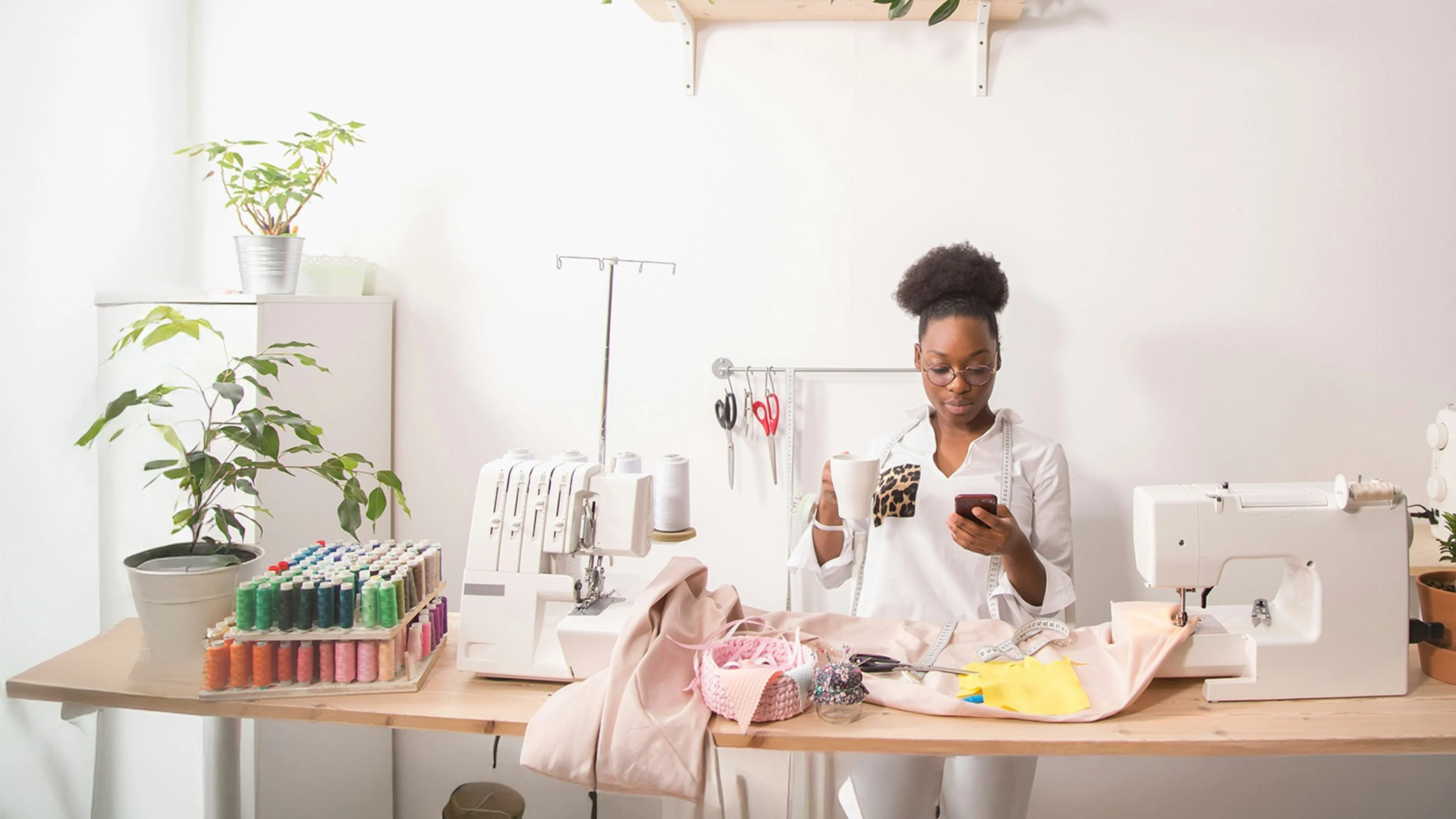 Woman using a sewing machine while holding a phone, representing a small business owner managing operations with online banking tools.
