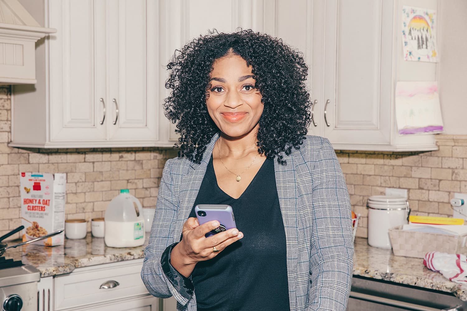 Woman in chic blazer standing in kitchen holding her phone and smiling