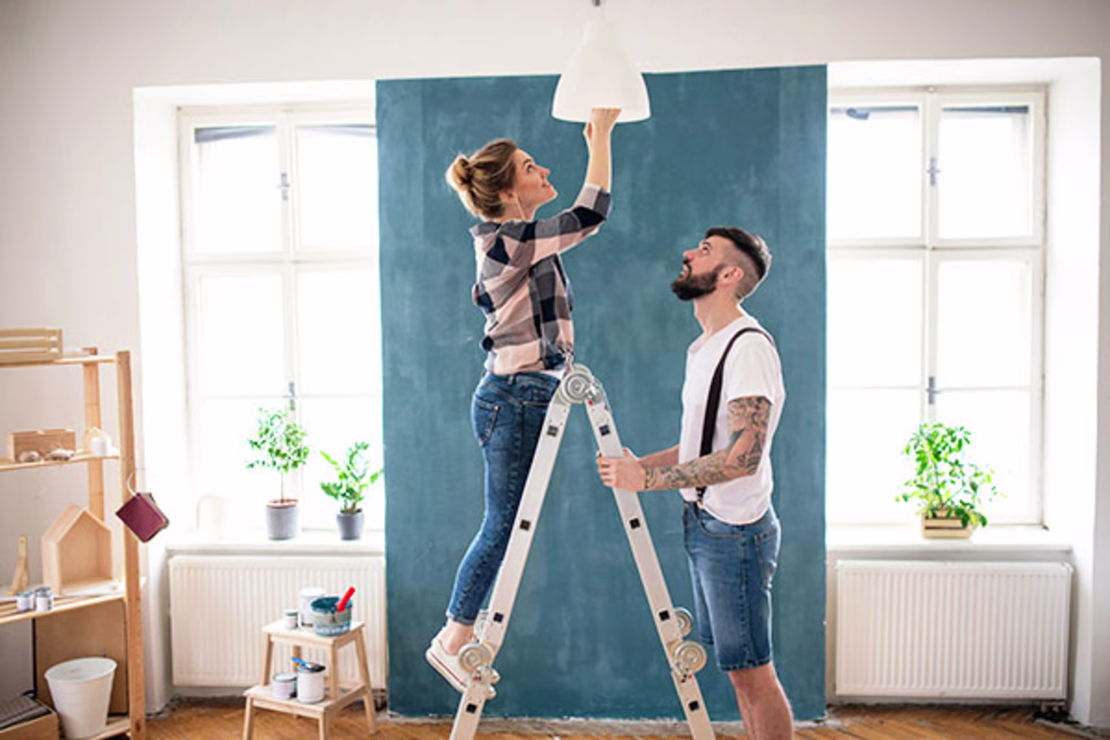 Couple working together to screw in a lightbulb, with the woman on a ladder and the man supporting it, showcasing teamwork in home improvement projects—ideal for discussions about home renovations, construction loans, and home improvement financing.