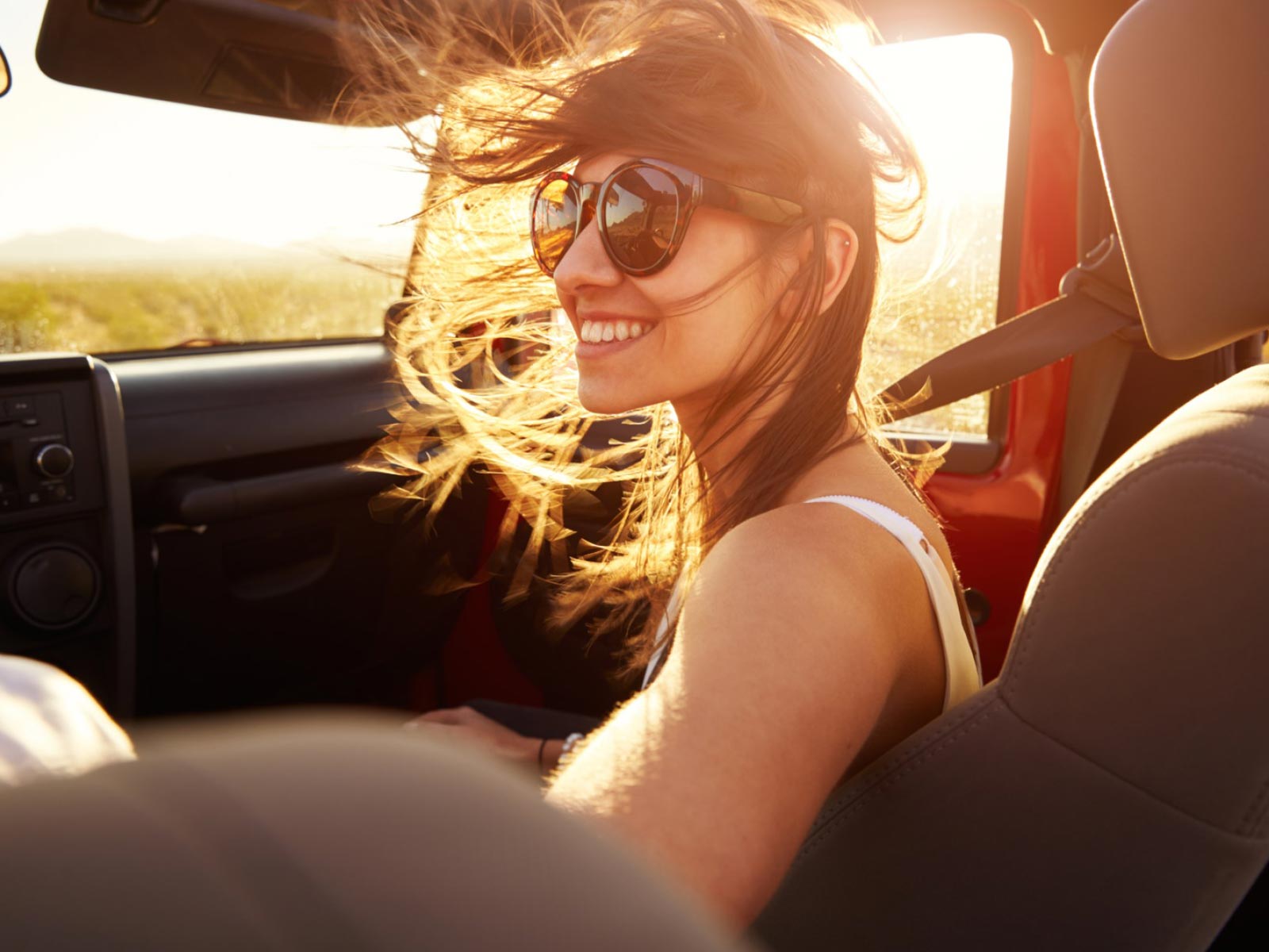 Young lady enjoying herself, wearing sunglasses, with the wind in her hair, while she's riding as a passenger in a convertible all-terrain vehicle, with the sun setting behind her.