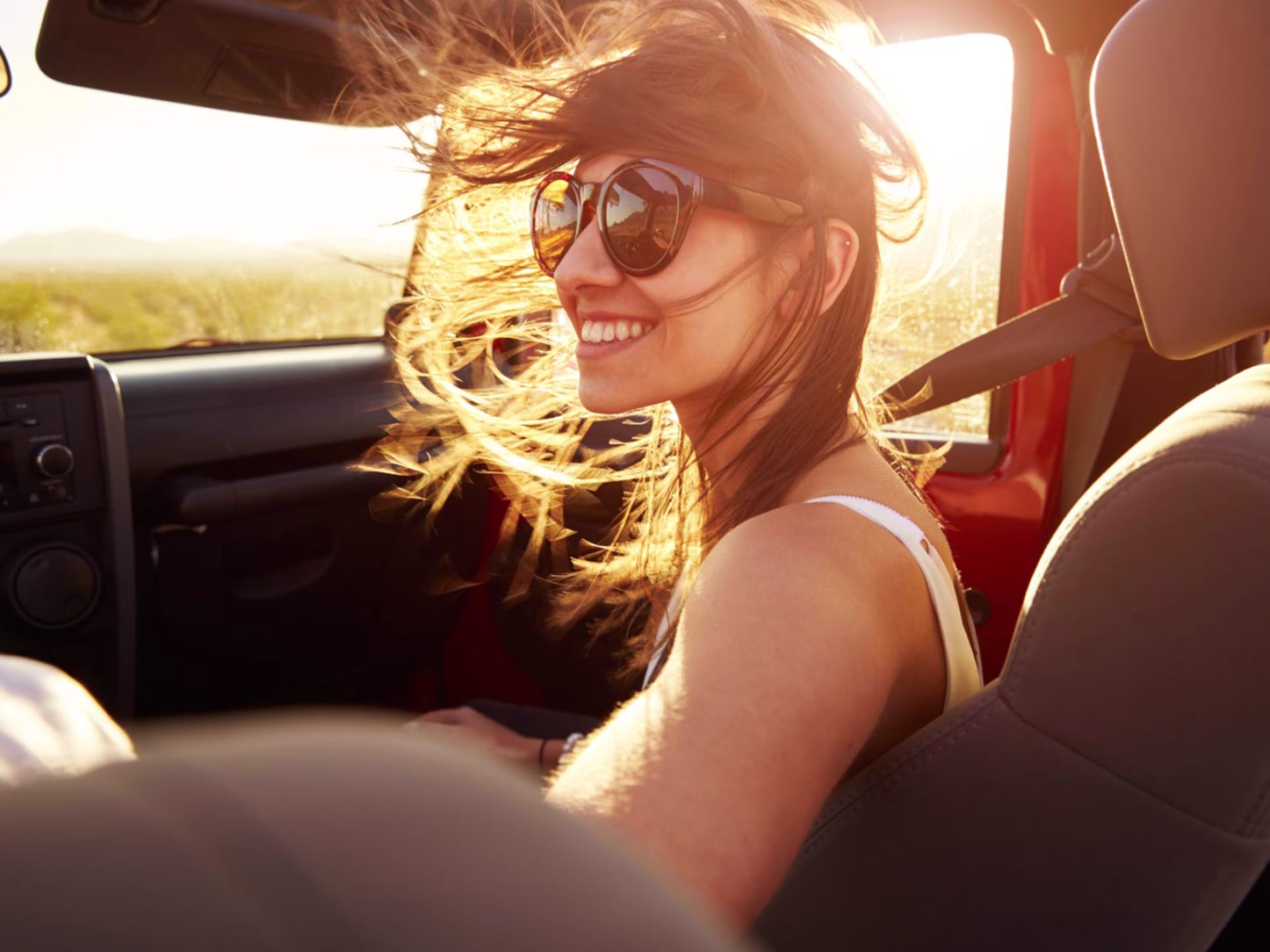 Young lady enjoying herself, wearing sunglasses, with the wind in her hair, while she's riding as a passenger in a convertible all-terrain vehicle, with the sun setting behind her.