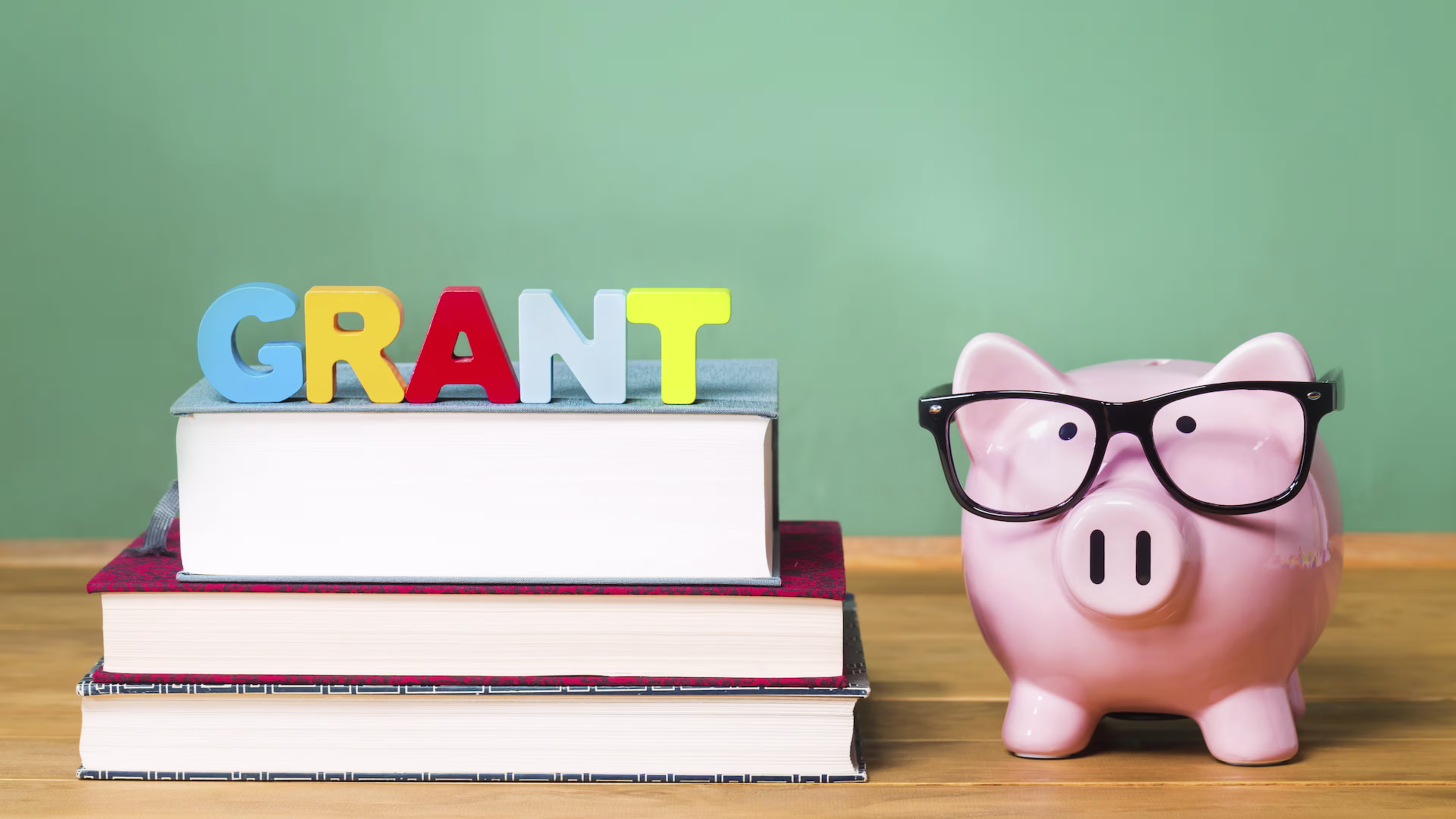 The word grant is spelled out in children's letters on top of schools books and a piggy bank with eyeglasses next to it.