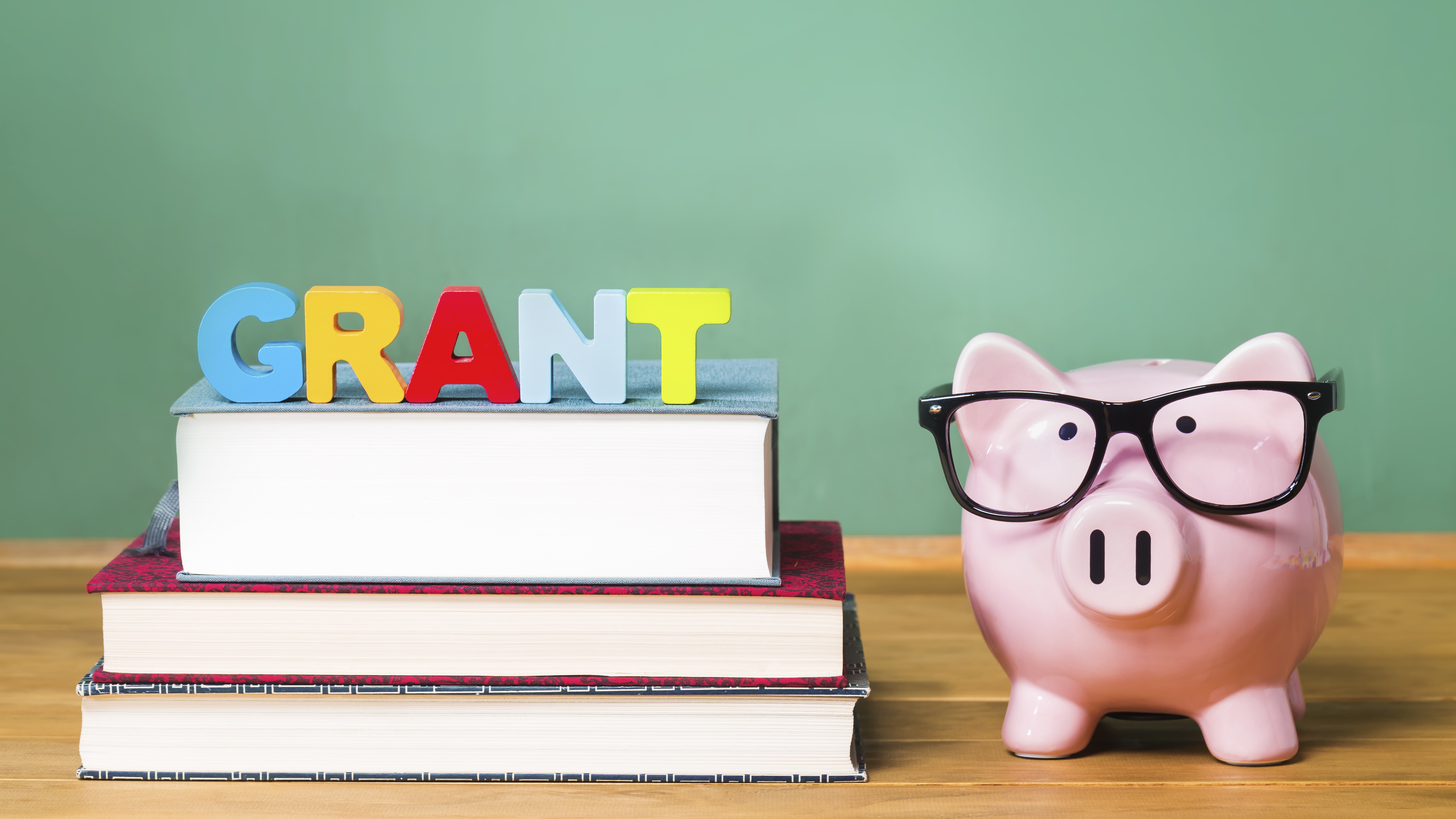 The word grant is spelled out in children's letters on top of schools books and a piggy bank with eyeglasses next to it.
