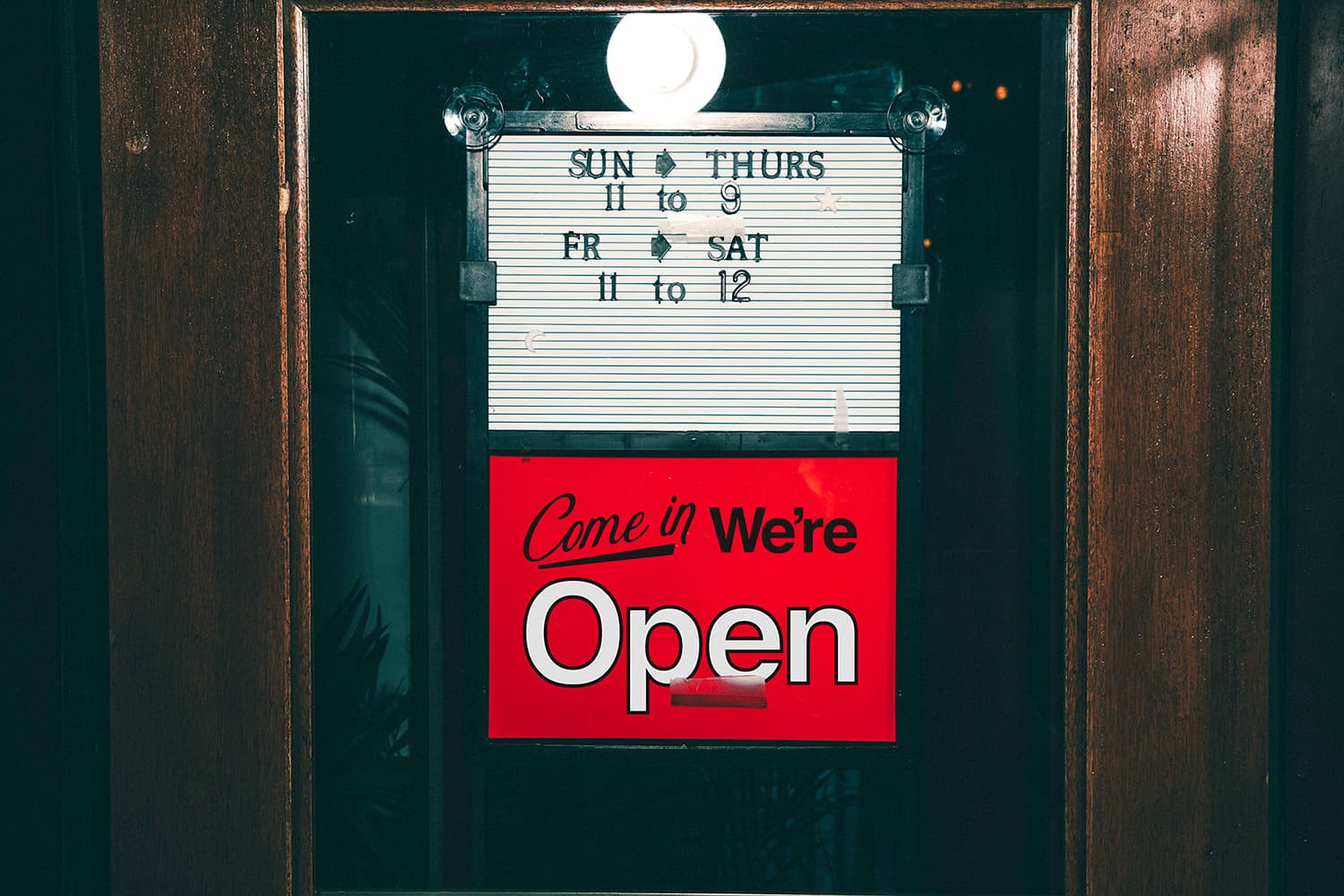 Wood-framed glass door with hours and "Come in we're open" signage