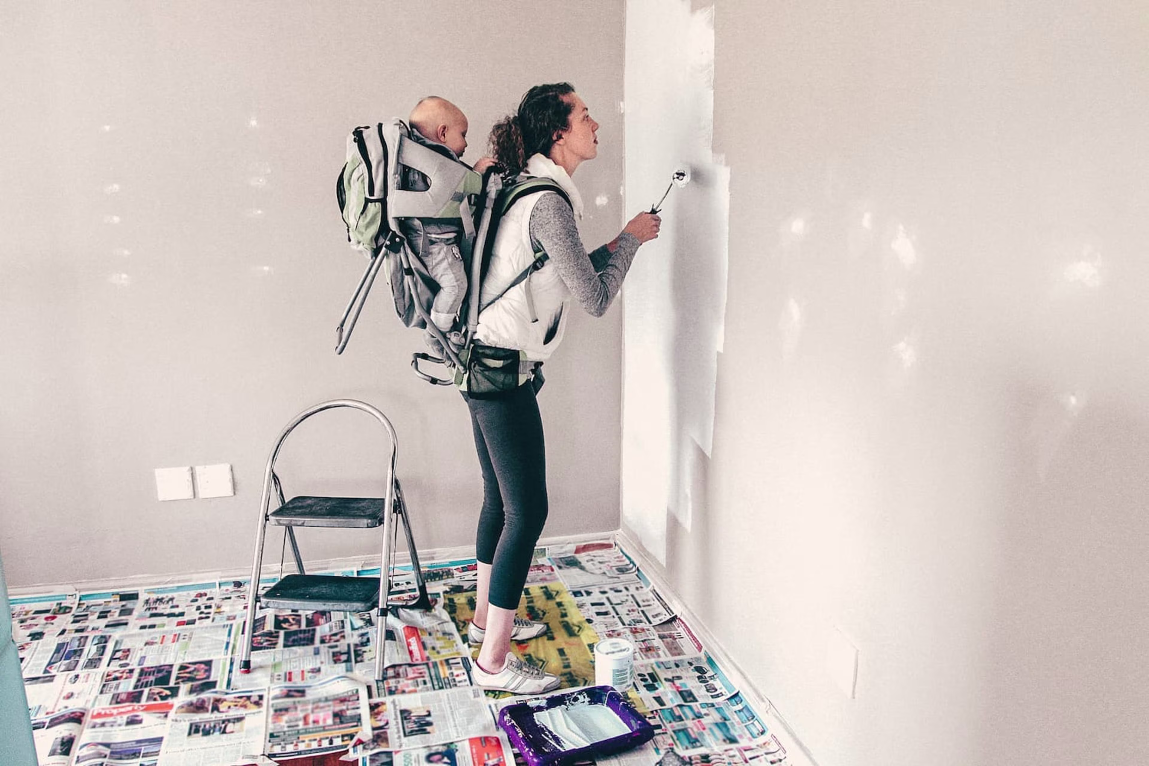 Woman paints a wall with child in baby carrier on her back.
