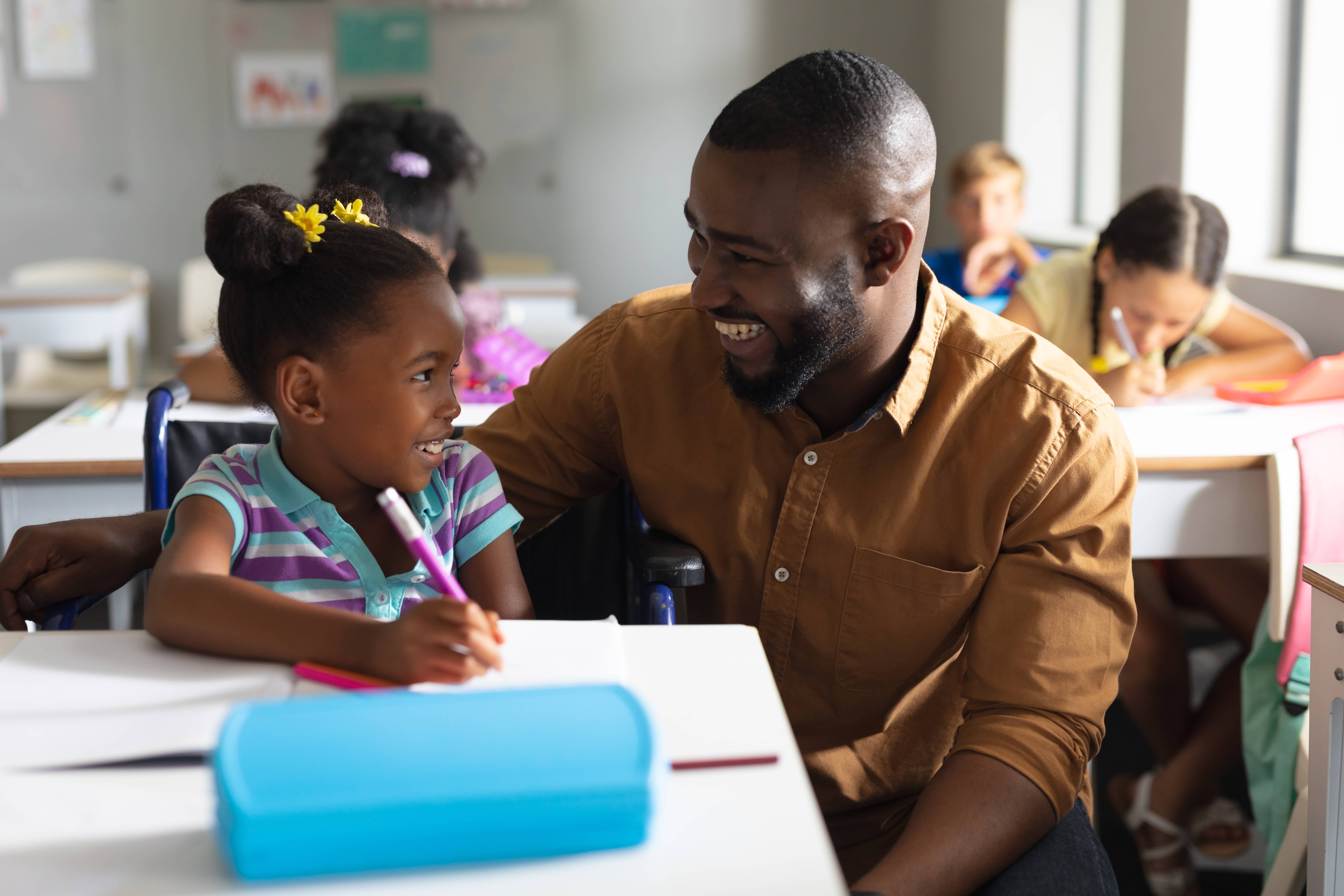 Male teacher smiling with a young student at her desk in a classroom