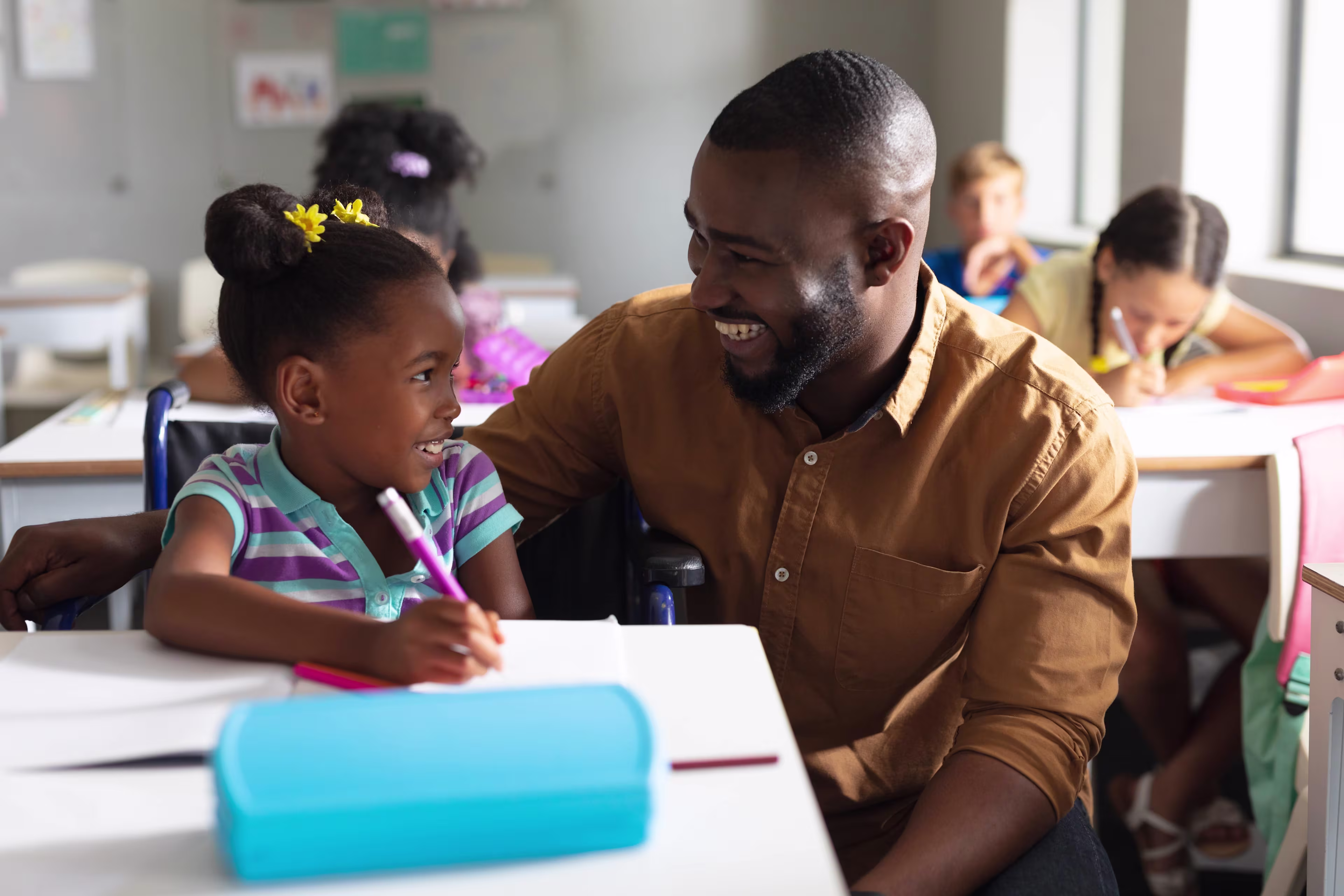 Male teacher smiling with a young student at her desk in a classroom