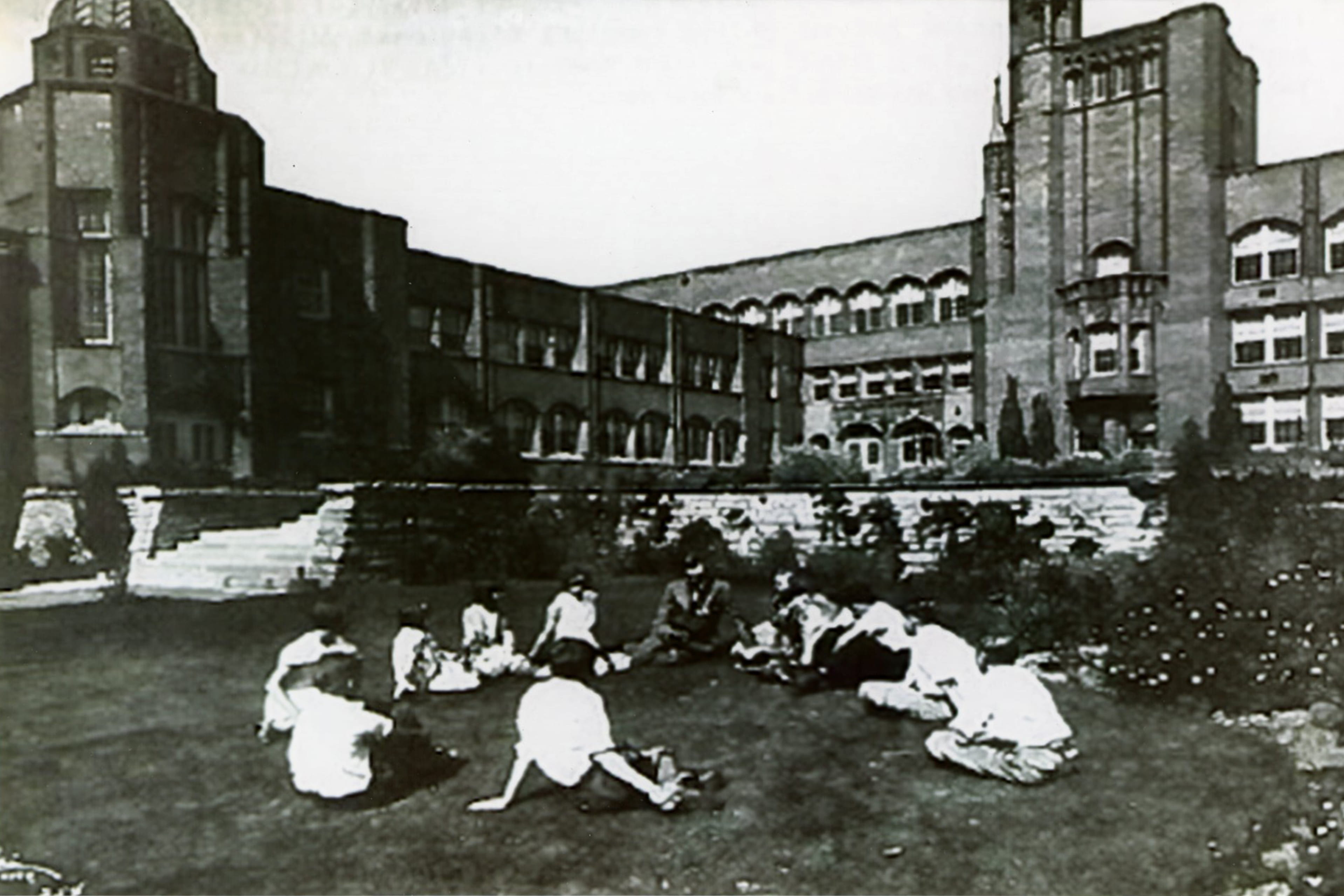Old black and white photo of people sitting in the grass in front of a building