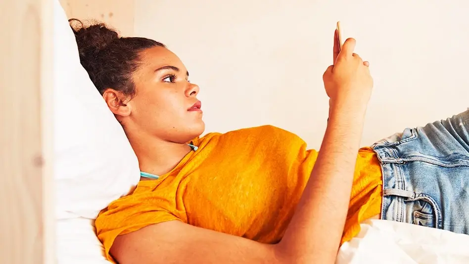 A woman in an orange shirt laying down and scrolling on a mobile phone.