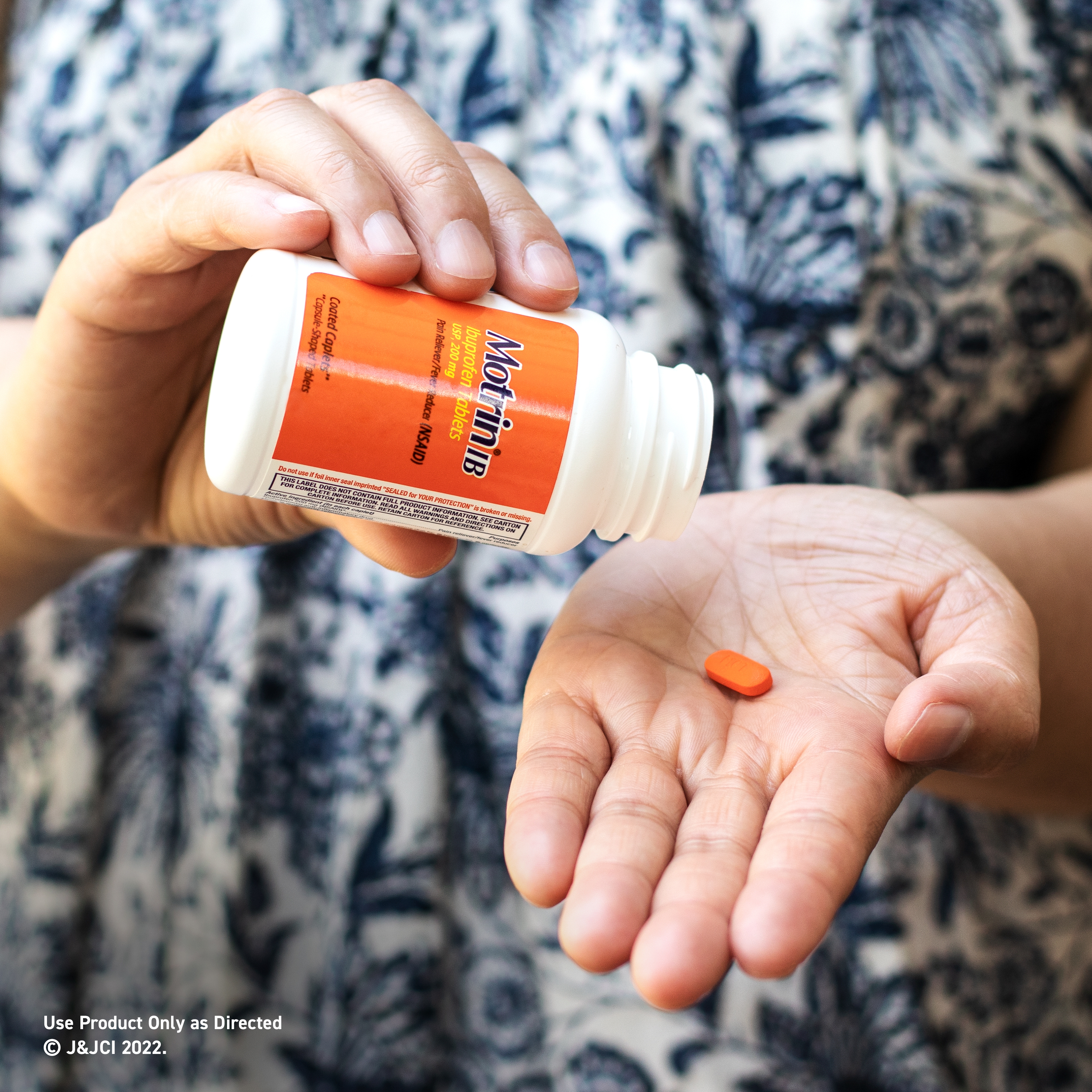 Close-up of a person's hand pouring a single orange Motrin IB tablet from a bottle onto their palm. The background is out of focus, showing a patterned shirt.