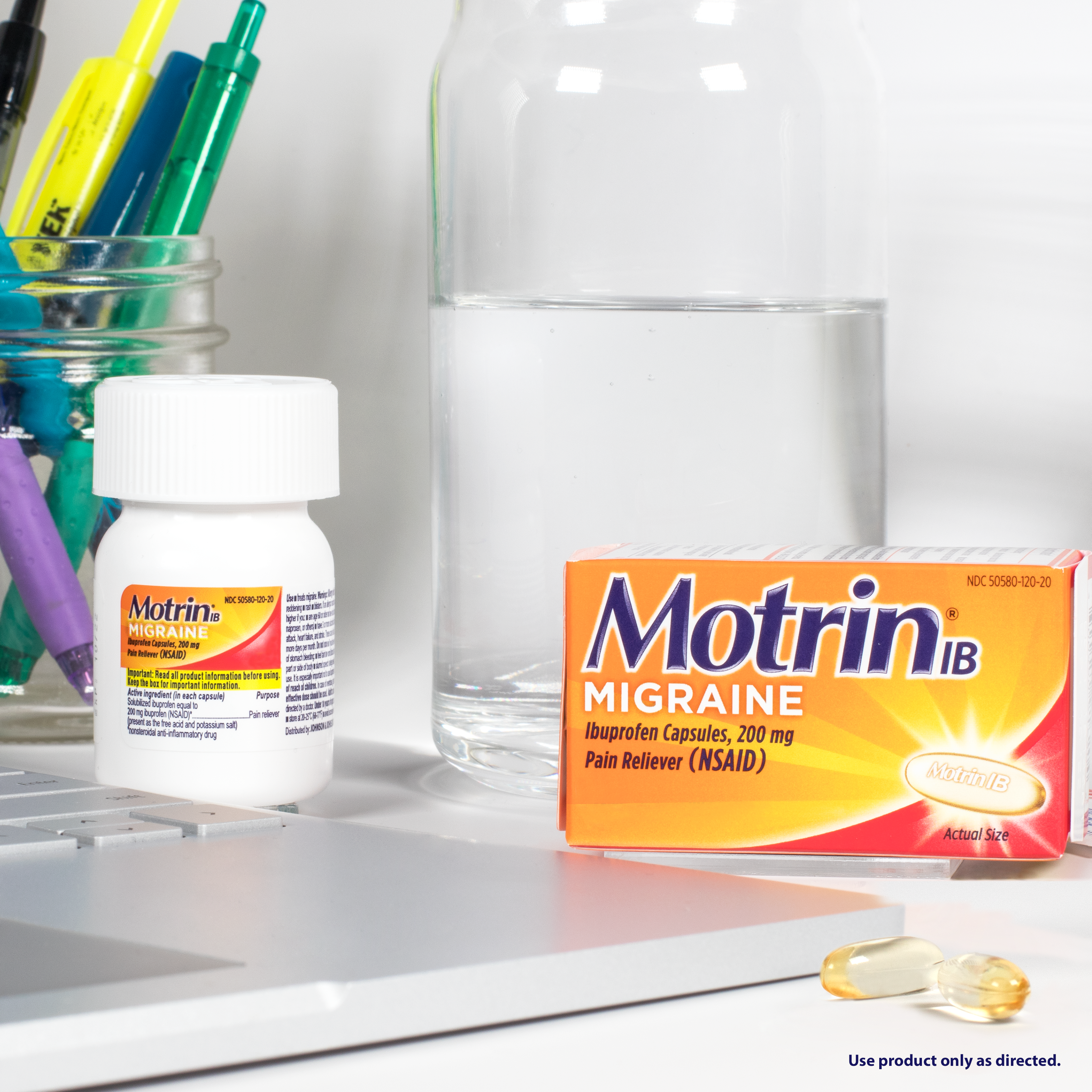 A box of Motrin Migraine pain reliever, a bottle of the same medication, two capsules, a glass of water, and office supplies near a laptop on a desk.