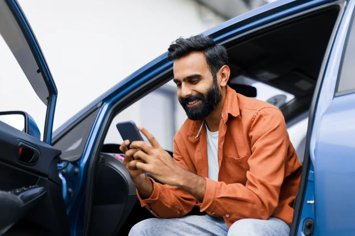 Positieve emotie - man kijkt naar telefoon en zit in deuropening van auto