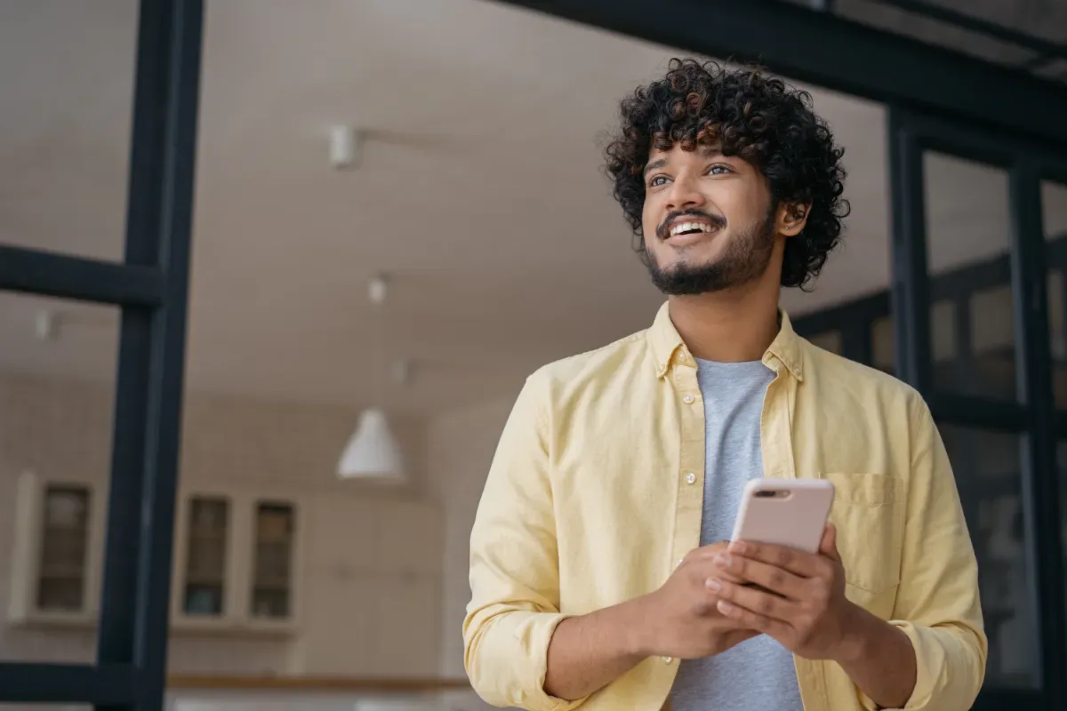 Positieve emotie - man in woonkamer kijkt vrolijk weg met telefoon in hand