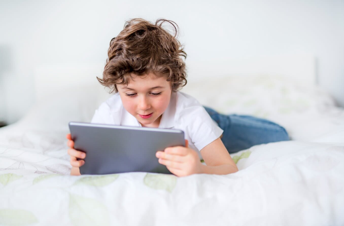 Un bambino che fissa lo schermo di un tablet mentre è a letto.