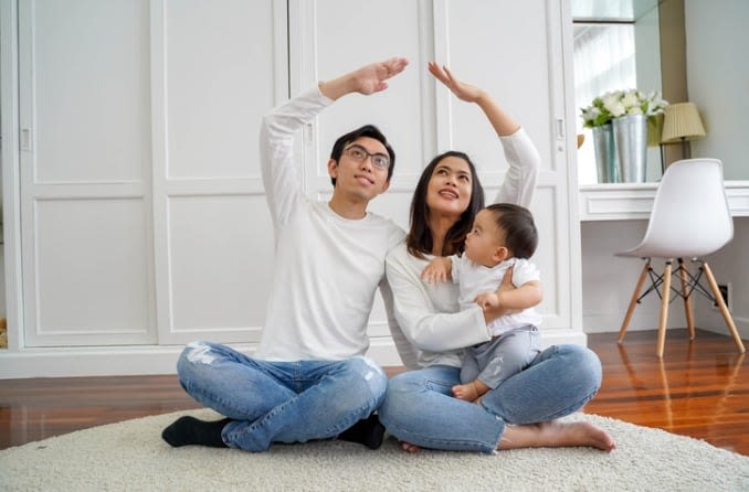 Young family with baby poses on living room floor, holding their arms up in the shape of a roof.