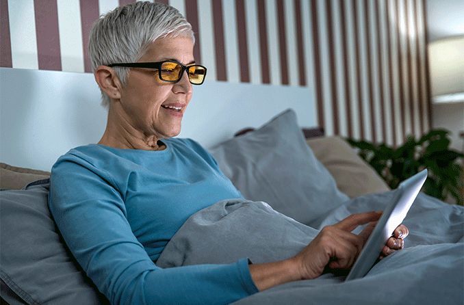 woman in bed on her computer tablet wearing blue light blocking glasses